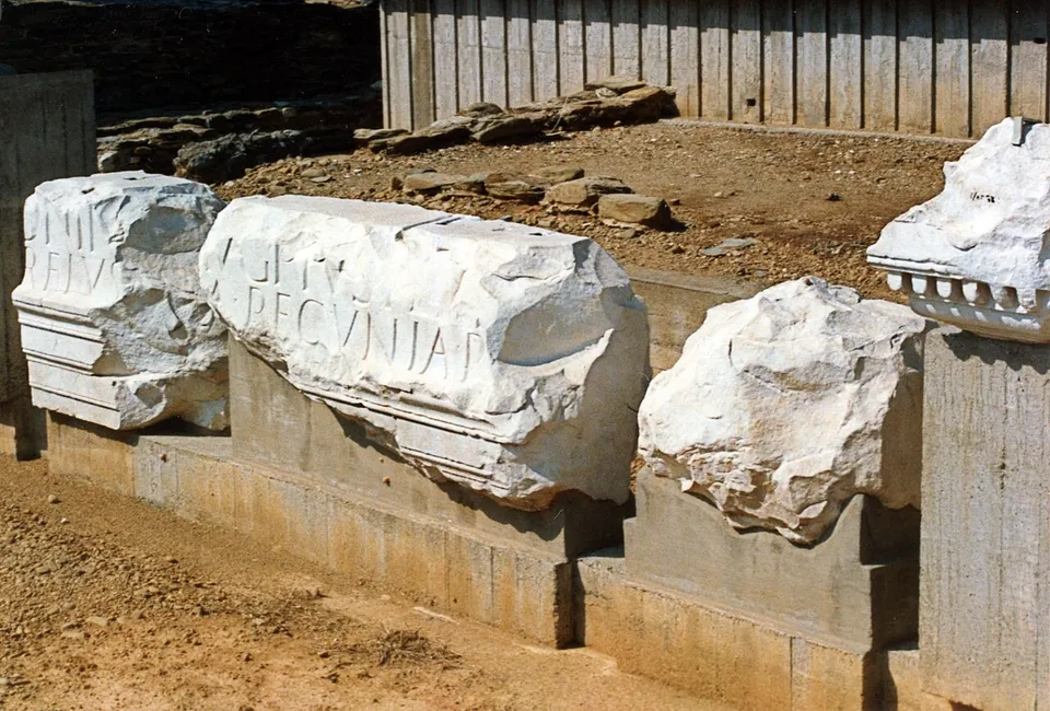Three broken blocks of white marble set on a concrete base in the open, two of them carrying large carved Roman capitals, with an excavation and a wooden fence behind.