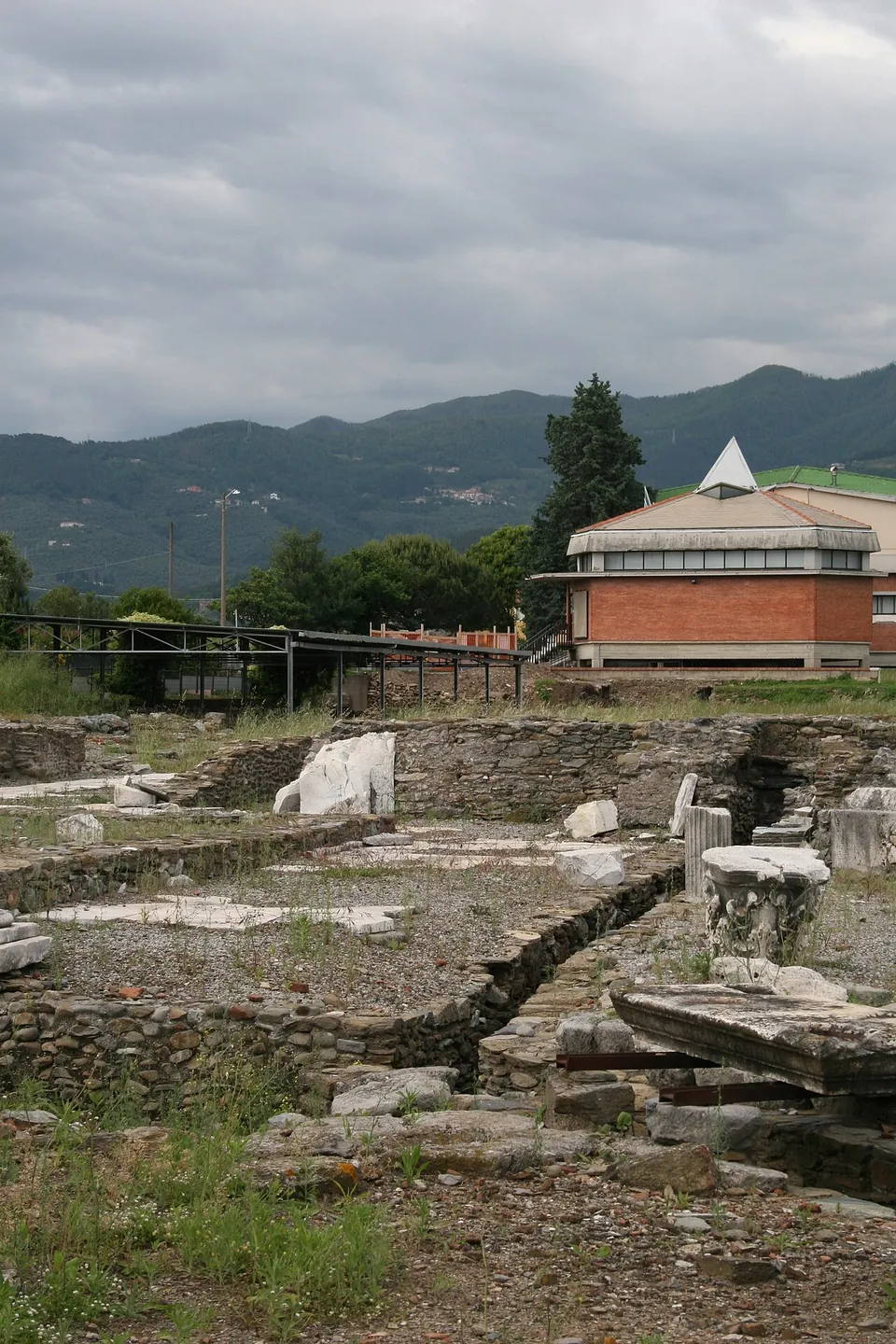 Excavated foundations with broken column drums and white marble blocks in the foreground, and behind them a modern brick building with a white pyramid-shaped roof light, against wooded hills.