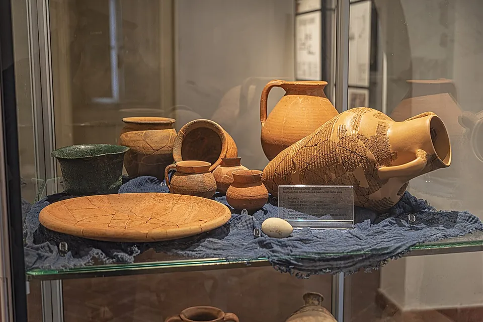 A glass display case holding pottery, jugs, bowls, a broad plate and a large amphora, laid on a blue cloth.