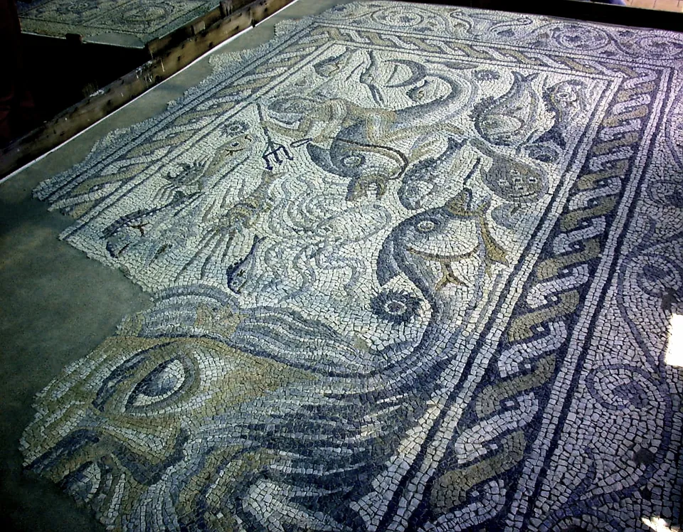 A mosaic floor of small black, white and grey stones showing fish and other sea creatures inside a twisted-rope border, with larger curving shapes along its lower edge.