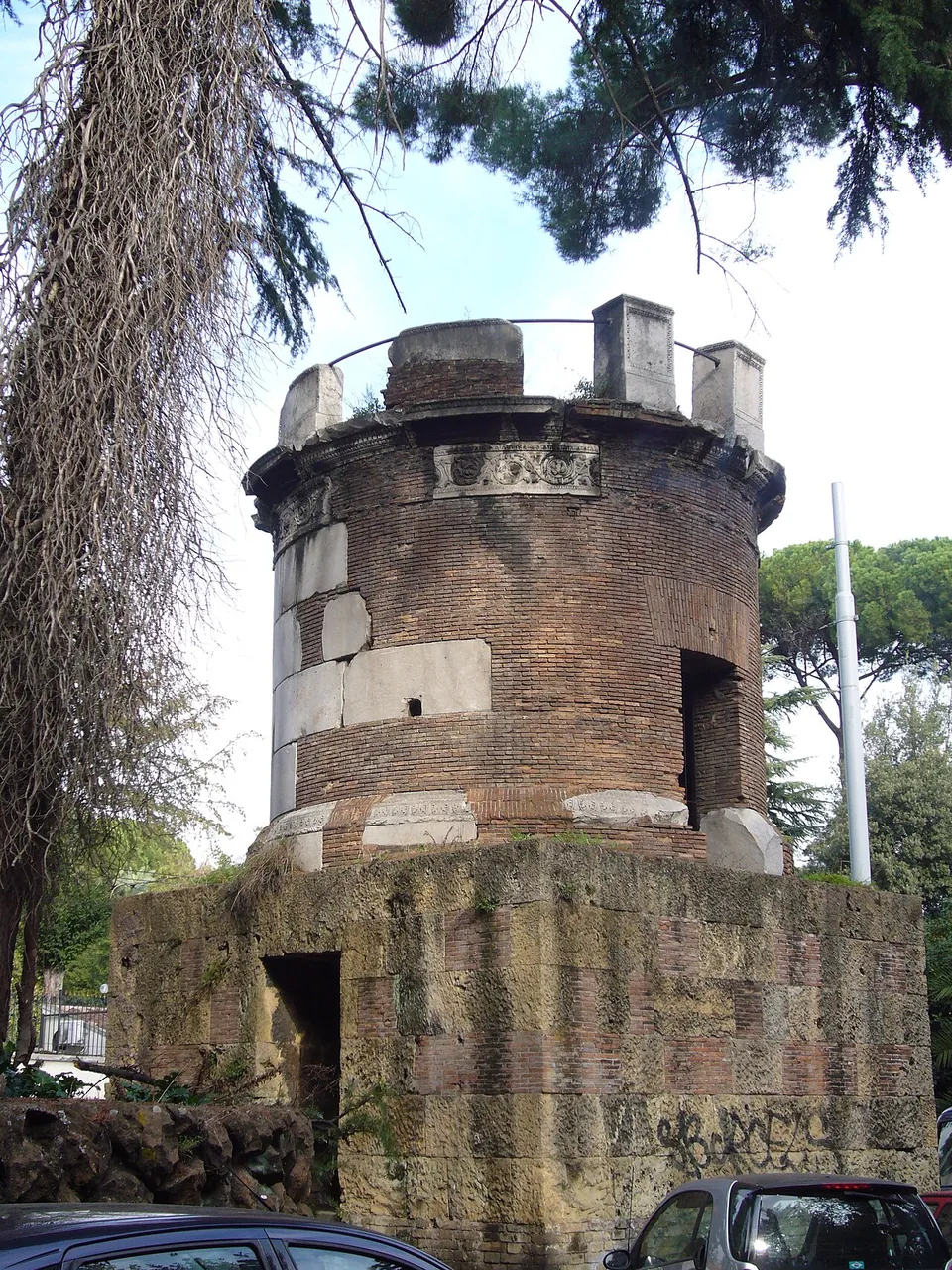 A round brick tomb set with blocks of white marble and a carved frieze below its rim, standing on a squared base beside a road, under trees, with parked cars in front.