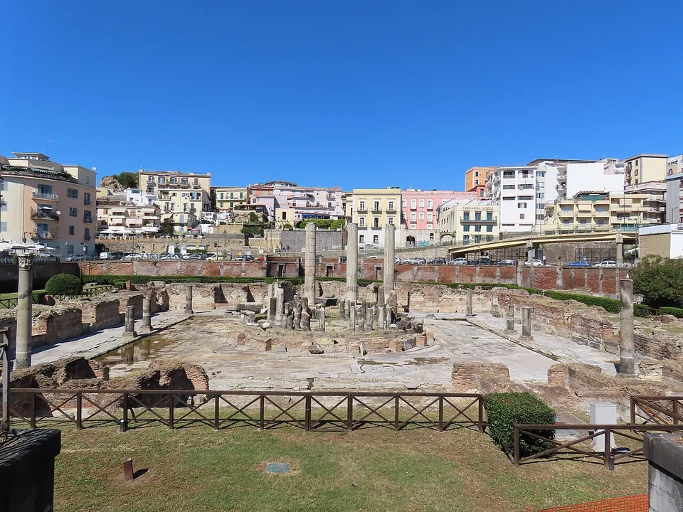 A broad square courtyard paved with pale stone slabs, with a low round podium at its centre ringed by broken column stumps and, behind it, three tall plain columns; a wooden fence and grass in the foreground, and pastel apartment blocks above a brick boundary wall beyond.