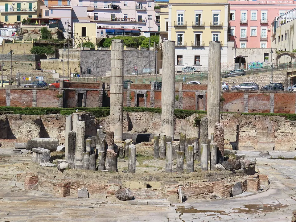 A cluster of broken column shafts of different heights standing on a round brick podium, with three much taller columns rising behind it in front of a brick wall, and parked cars and apartment buildings at street level above.