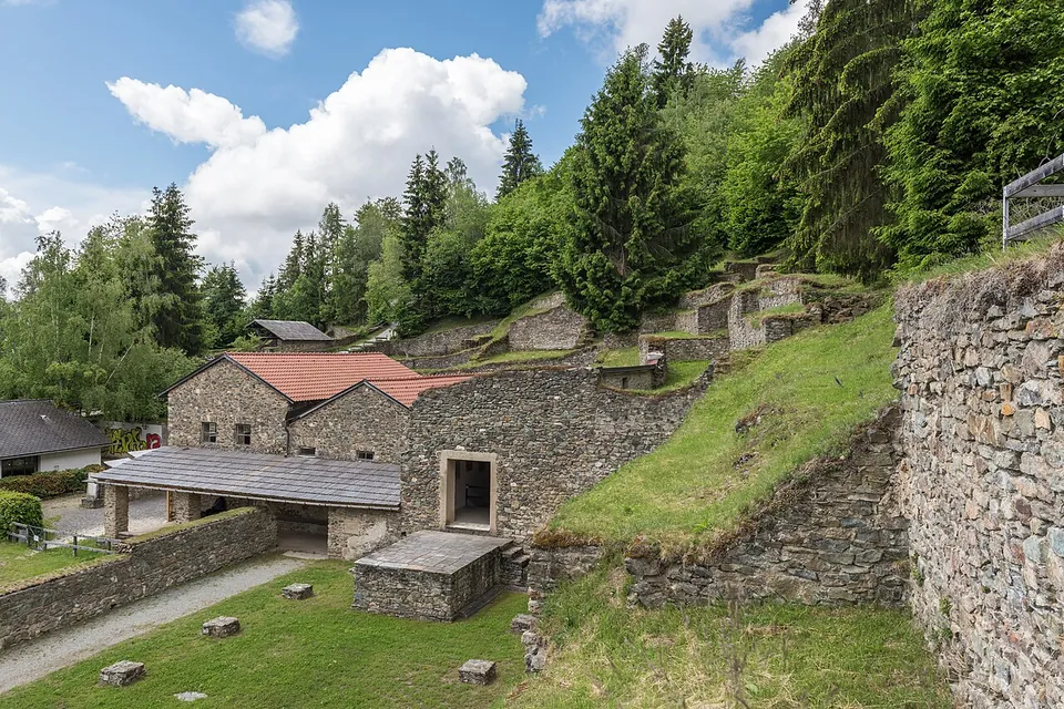 Stone buildings with red tiled roofs and a covered walkway among excavated walls on a grassy slope, with conifers and a blue sky with white clouds behind.