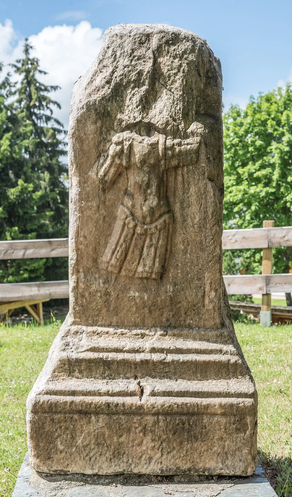 A weathered stone grave altar carved with a standing figure in a niche, set on a plinth in the open in front of a wooden fence and trees.