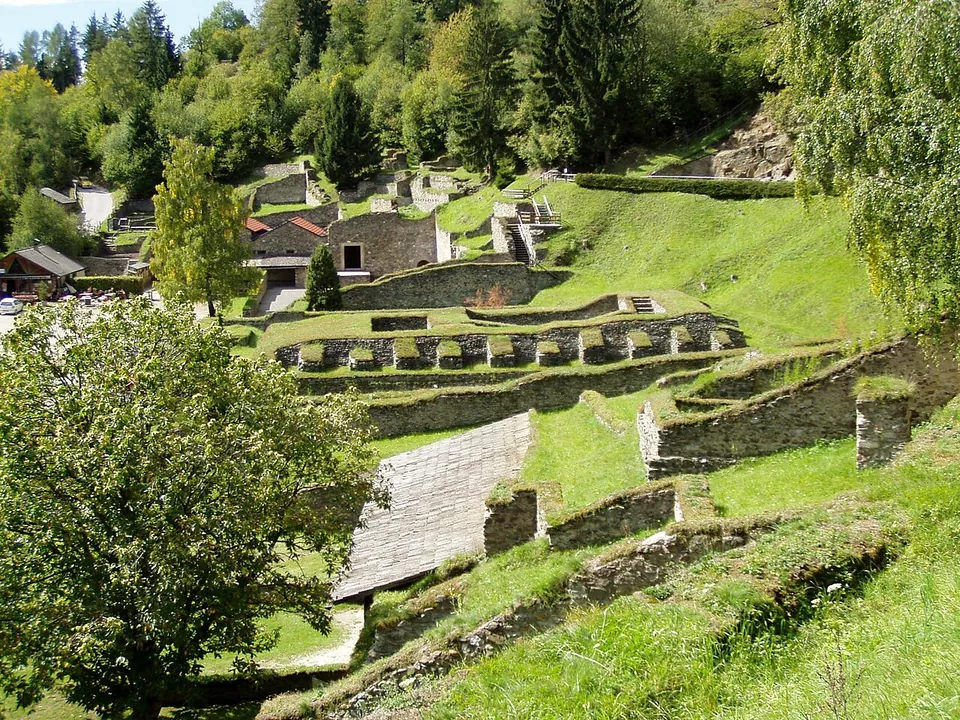 A green hillside terraced with the low stone walls of excavated buildings, some under modern roofs, rising to a small group of rebuilt houses and woodland at the top.
