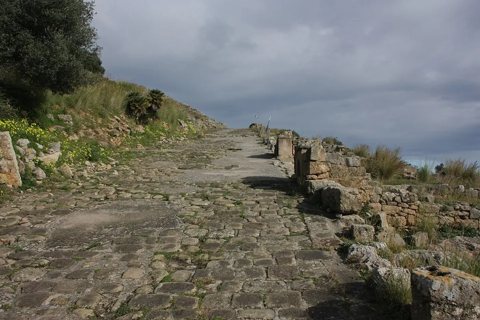 A broad ancient street paved with irregular stones running straight away from the camera between low ruined walls, under a grey sky.