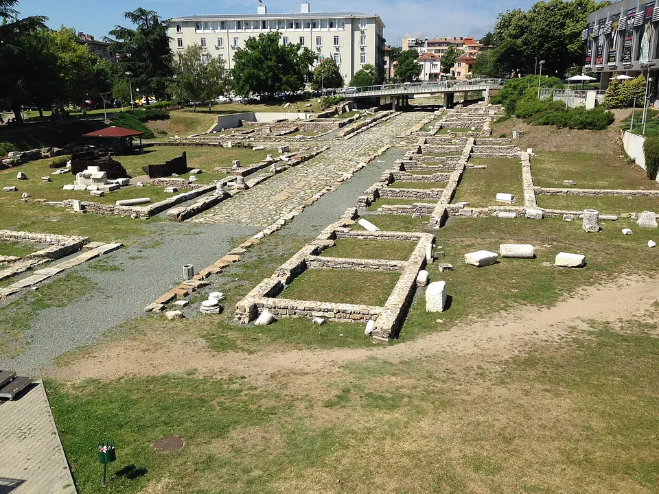 An excavated area seen from above, a gravel-and-stone street running through low rectangular stone foundations set in mown grass, with scattered column drums and blocks, trees and modern buildings beyond.