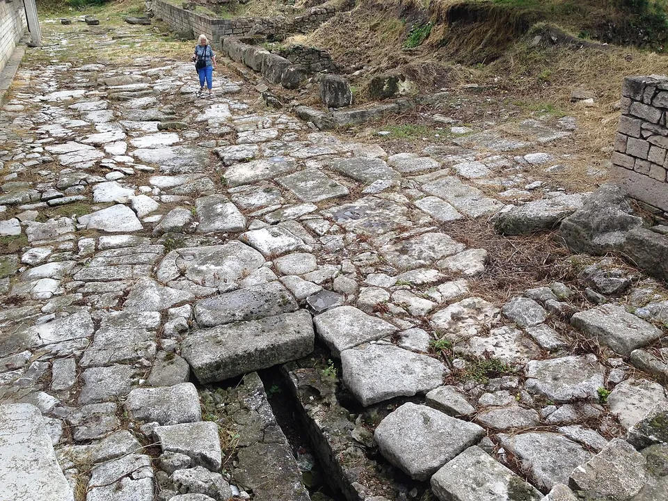 A Roman street of large worn paving slabs with a drain channel down one side, low walls along it and a visitor walking in the distance.