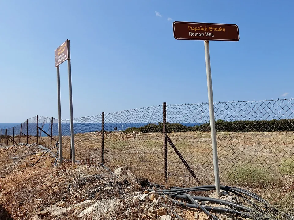 Two brown signposts in Greek and English, one reading "Roman Villa", beside a chain-link fence around a dry field by the sea.