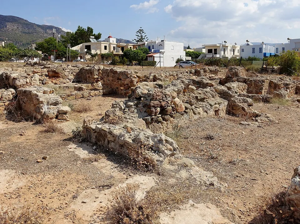 Low ruined walls of rough stone across a dry, scrubby excavation, with white holiday houses, trees and a mountain behind.