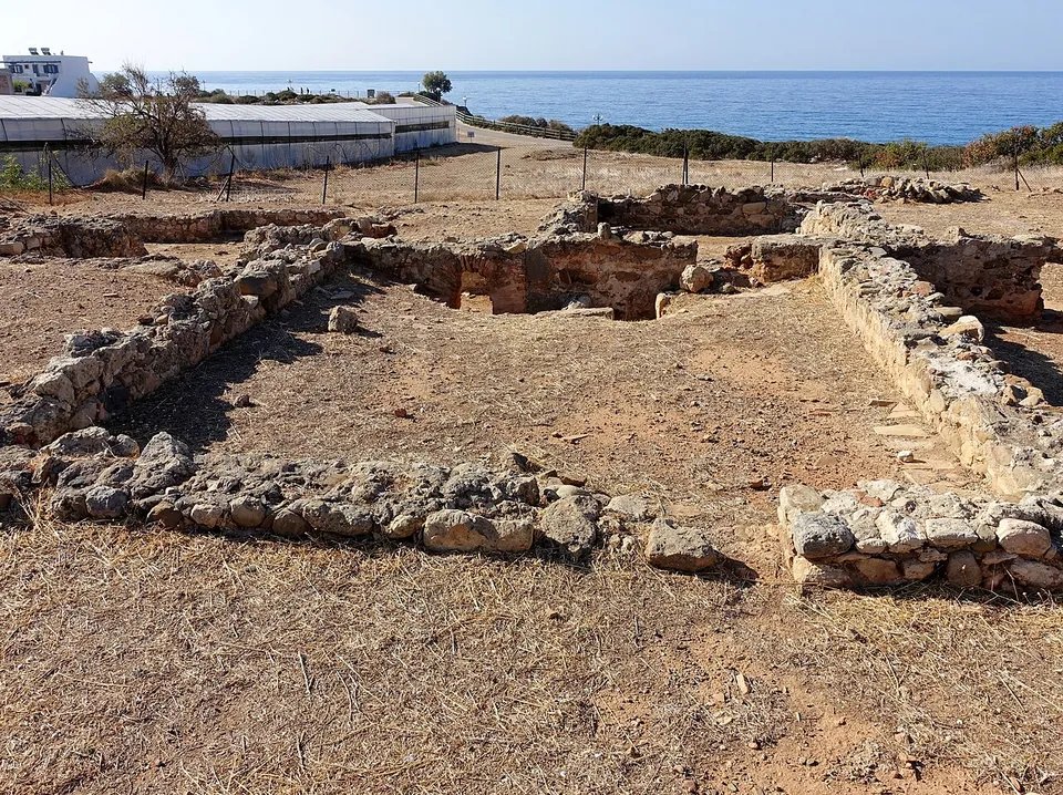 The low stone walls of rectangular rooms on bare ground, with greenhouses, a fence and the sea behind.