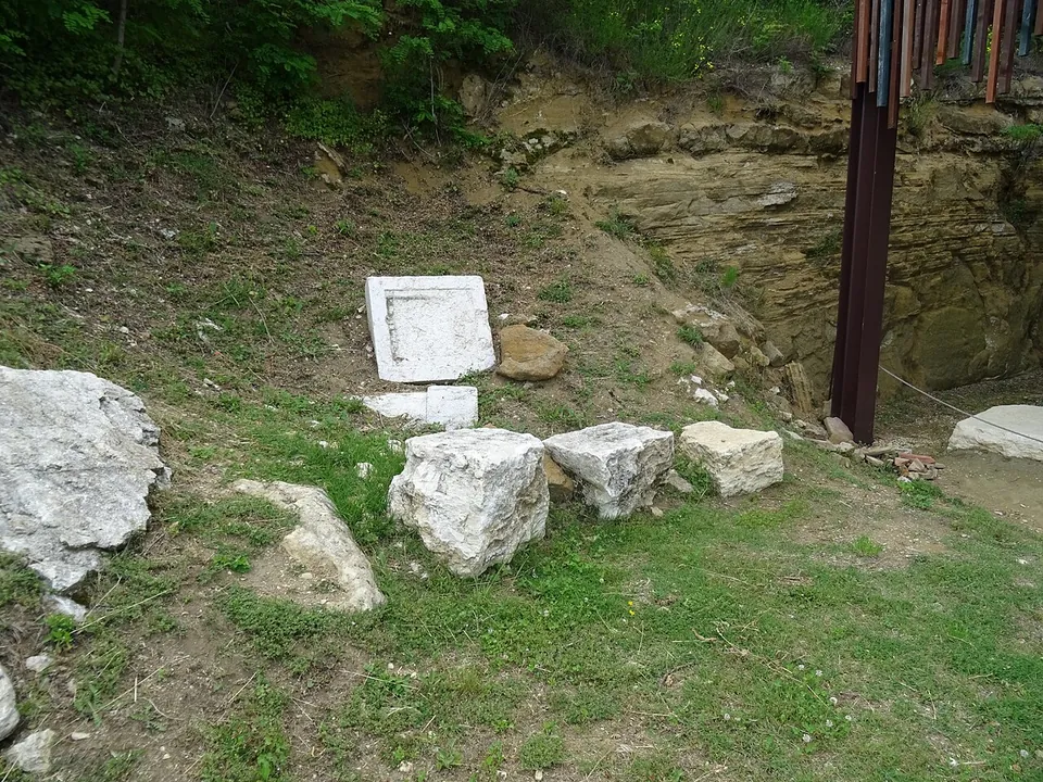 A grassy hillside terrace with a gravel path leading to a large open shelter of dark steel posts, its roof edged with a screen of vertical wooden slats, standing against a wooded rock face; low excavated walls lie beneath it and an information panel stands in front.