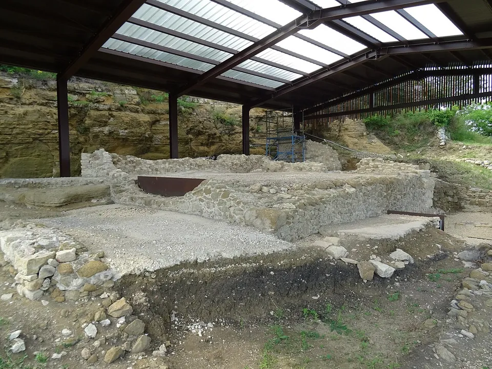 Under a translucent roof on steel posts, a raised rectangular platform of rubble-stone walls with a gravelled top, a rusted metal plate set into its face, and exposed rock strata behind.