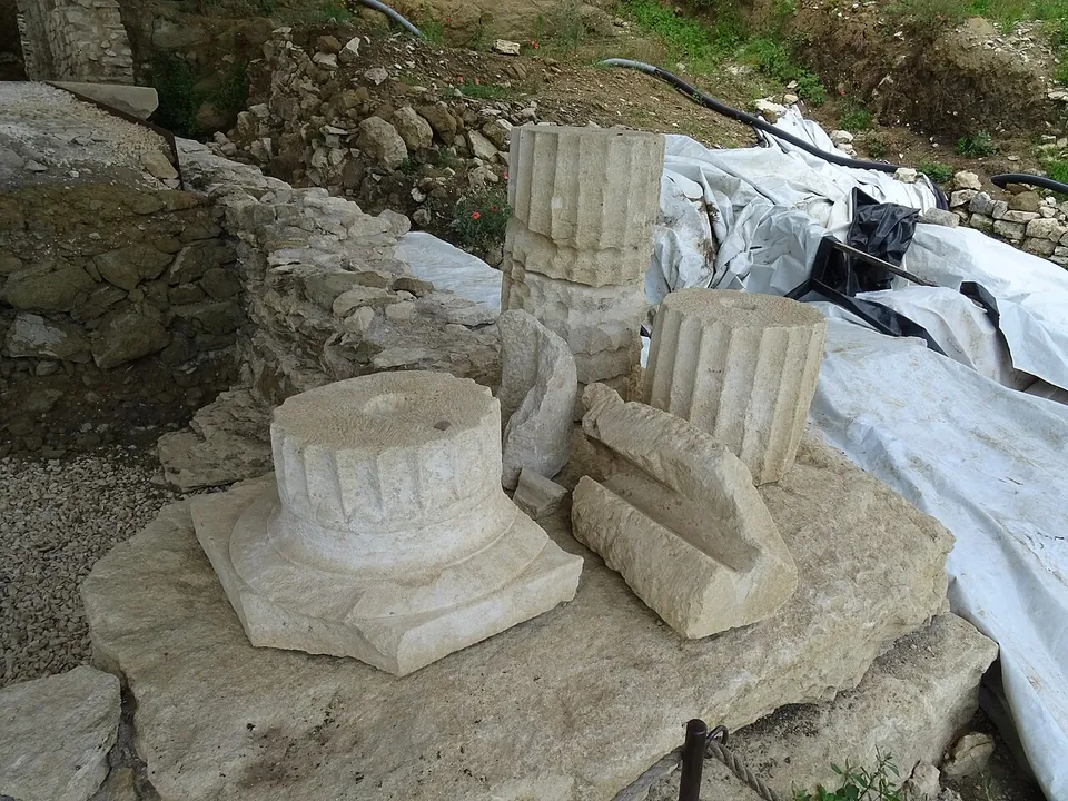 Two fluted column drums and a column base with a moulded plinth, with other worked stone fragments, set together on a stone block beside a rubble wall, with protective sheeting behind.