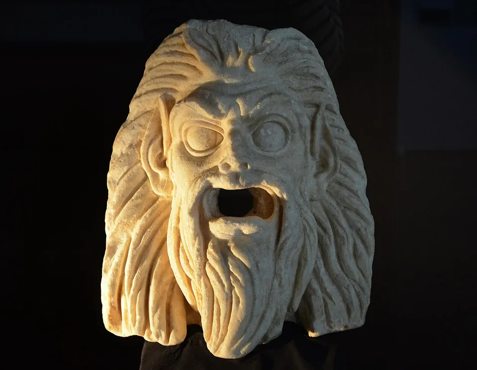 A white marble mask of a bearded face with wild hair, staring eyes and a gaping open mouth, lit against a dark background.