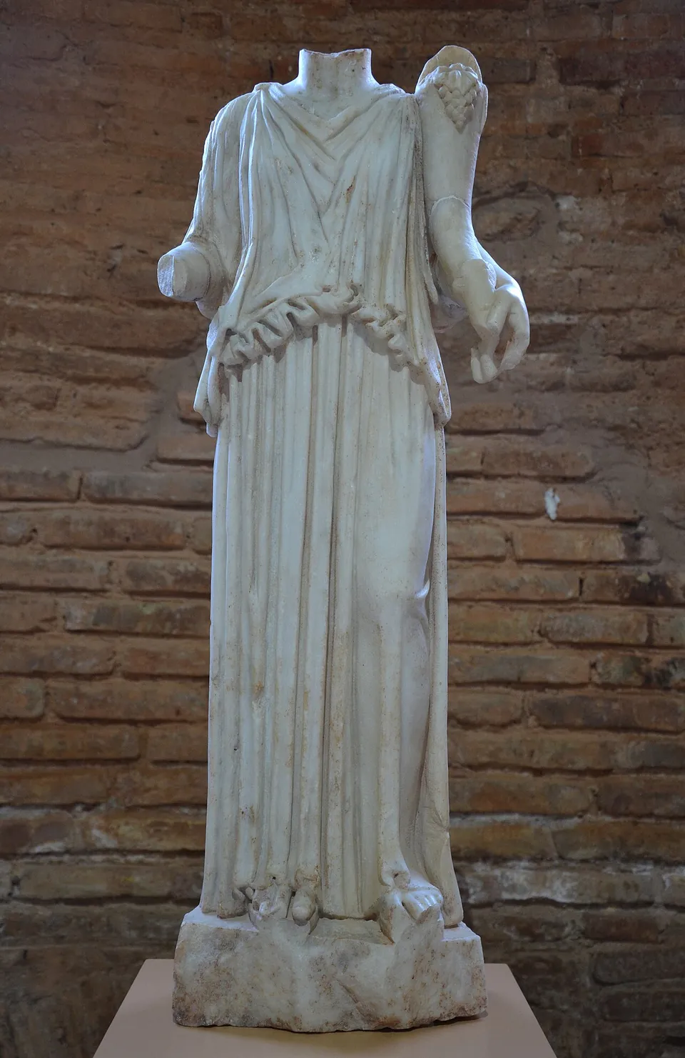 A small headless marble statue of a woman in a long draped robe, holding a horn of plenty in the crook of her arm, in a museum display.