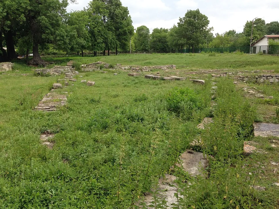 A grassy field with lines of low squared stone blocks marking out foundations, trees along the edge and a house behind a fence.