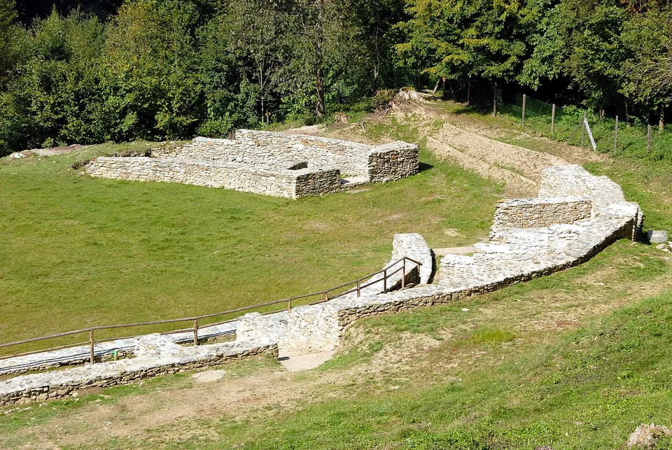 Low curving walls of grey stone around the edge of a grassy arena, with a squared stone block at one end and woodland behind.