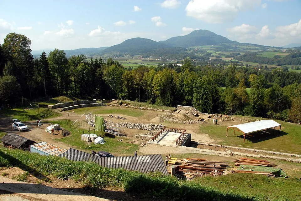 An excavation site in a clearing seen from a slope, with timber, sheds and a white canopy among exposed walls, and a wide farmed plain and rounded hills beyond.