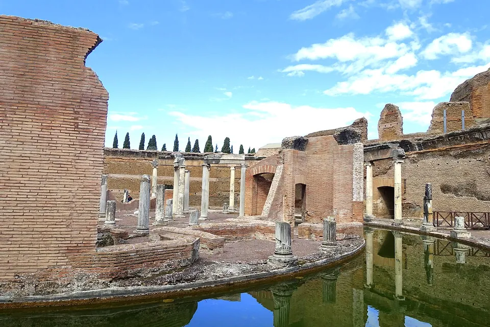 A ring of water curving round a low island of brick walls and broken column shafts, with taller ruined brick walls and a few standing columns beyond, under a blue sky.