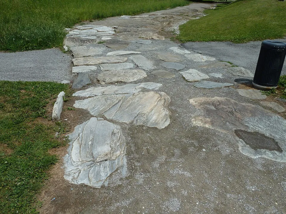 A path paved with large irregular flat stones running between strips of grass and asphalt, with a litter bin at the side.
