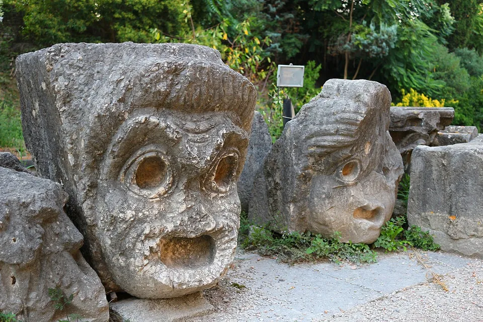 Two large blocks of weathered limestone carved as theatre masks, with round staring eyes and gaping open mouths, set on the ground among other carved stones.