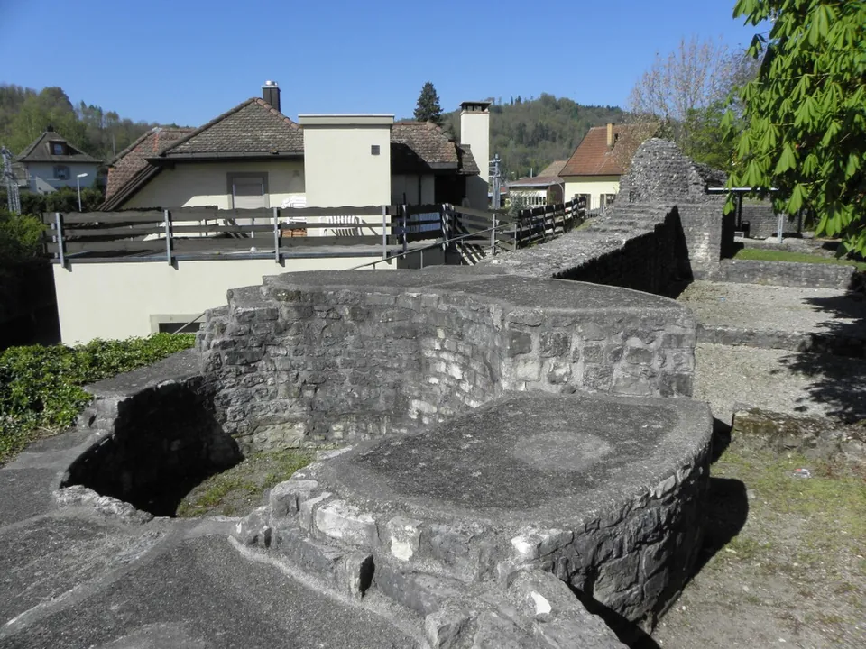 Rounded stone foundations of a tower with flattened, capped tops beside a stretch of wall, with houses and a wooden balcony behind.