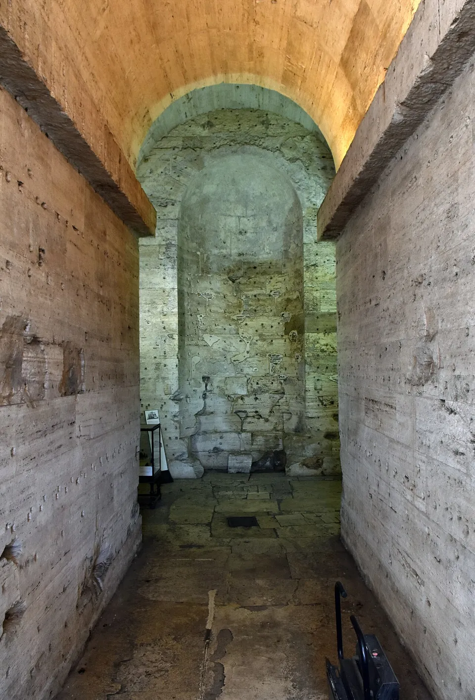 A long straight passage with walls and a vault of pale squared stone blocks pocked with small holes, ending in a tall recessed niche, over a floor of worn stone slabs.