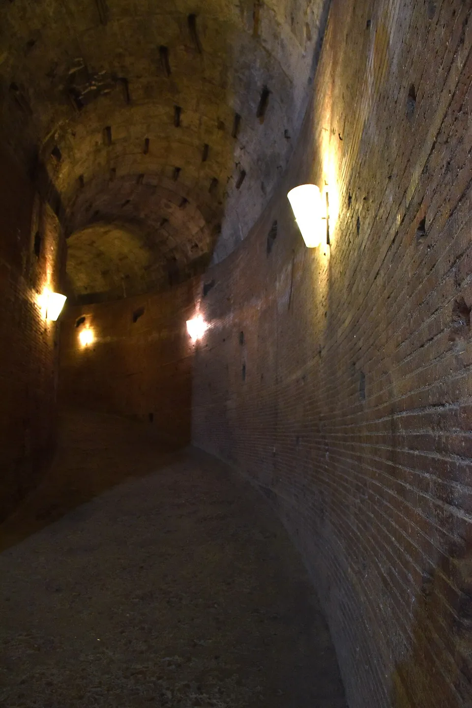 A brick-walled passage curving gently upward under a barrel vault, lit by lamps on the walls, its floor a smooth slope rather than steps.