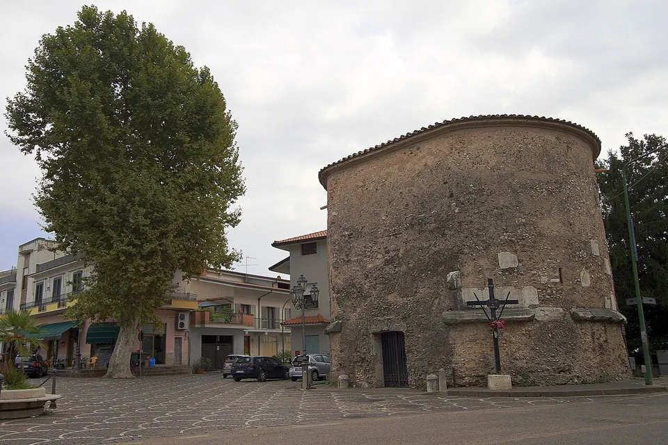 A squat round tower of rough grey stone under a low tiled roof, with a small doorway and a few squared stone blocks set into its face, standing in a paved square beside a large tree and modern houses.