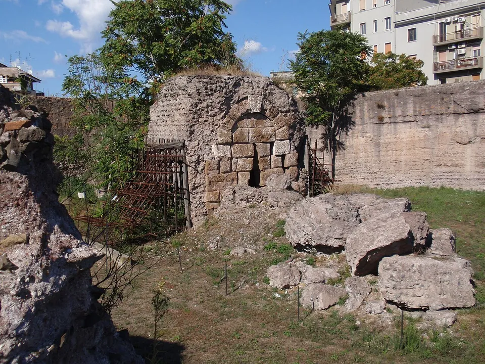 Inside the ring, a ruined block of squared stones with an arched opening, a rusted metal grille in front of it and large fallen blocks on the grass.