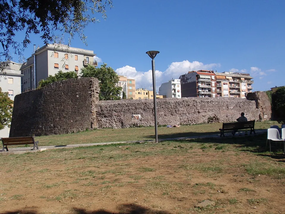 A park lawn with benches and a lamp post in front of a long, low curved wall of rough stone, with apartment blocks behind it.