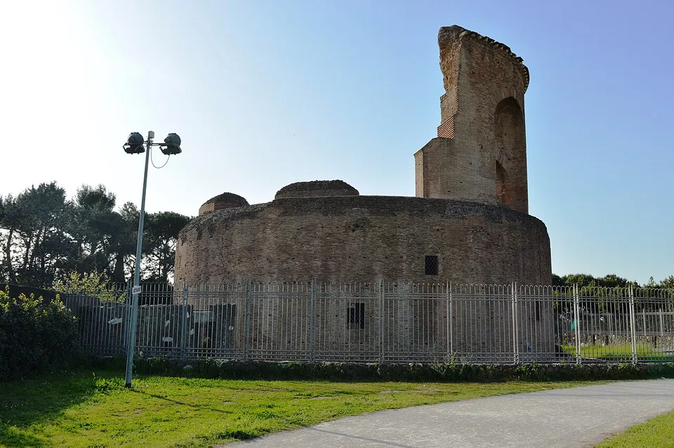 A round brick ruin standing behind a tall iron fence at the edge of a lawn, a fragment of its upper storey rising high above one side of the drum, with trees and a floodlight pole nearby.