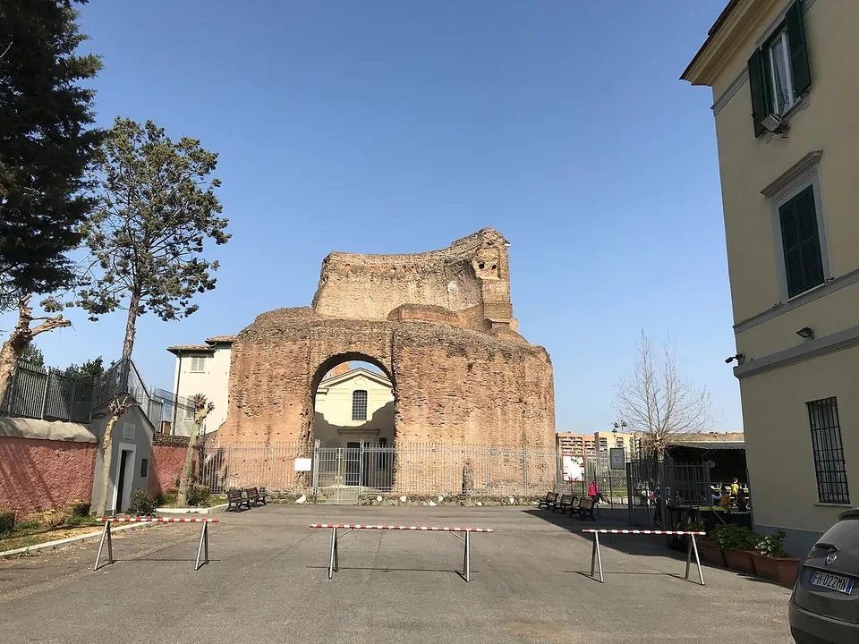 A paved forecourt with low barriers and benches in front of the ruined brick drum, through whose great arched opening the pale front of a much later building with a door and windows can be seen.