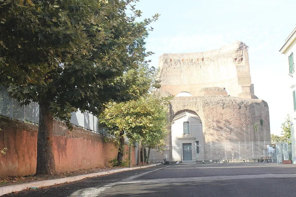 A paved lane between an orange garden wall and trees leading to the ruined brick mausoleum, with a small white doorway set into a great arch at its base.