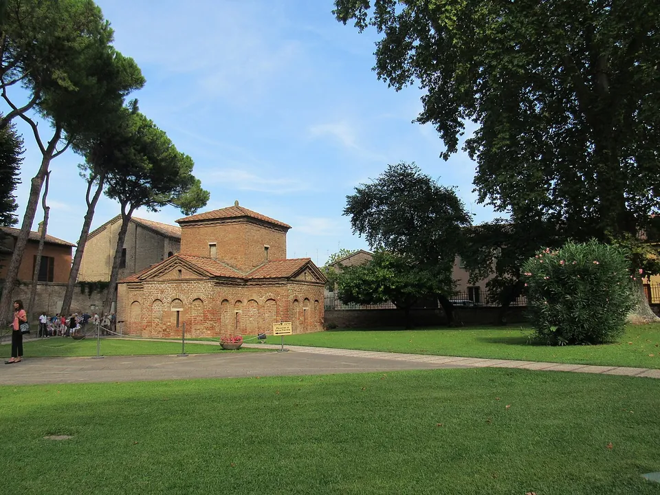 A small cross-shaped brick building with tiled gabled roofs and a square tower at its centre, its walls lined with shallow blind arches, standing on a lawn under tall pines and leafy trees, with visitors by a low building at the left.