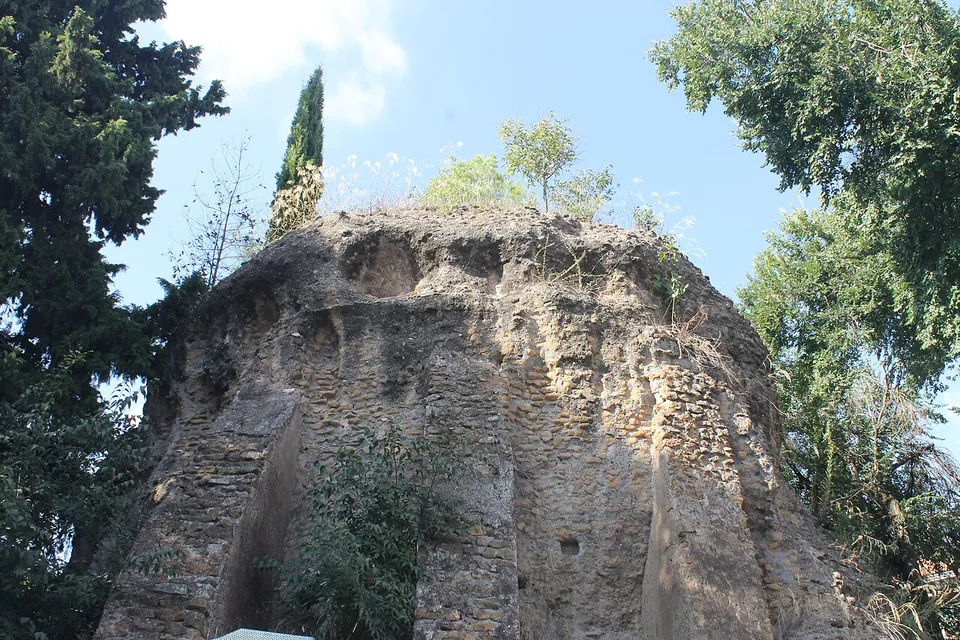 A squat, roughly round mass of weathered concrete and tuff, overgrown on top, standing beside a street among trees.