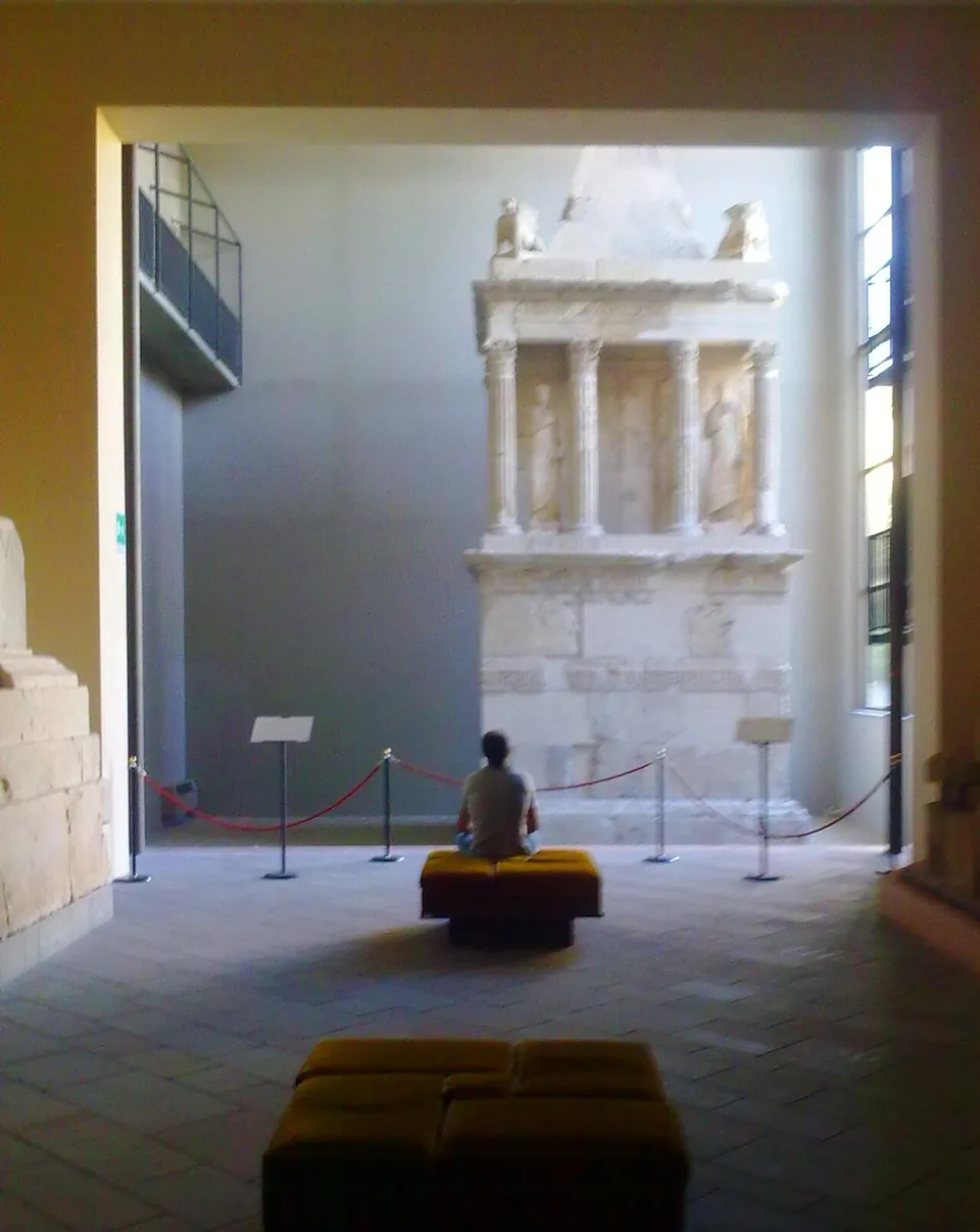 A tall pale stone funerary monument against the end wall of a high museum hall, with a square base, an open storey of columns with statues between them and a pointed roof; a visitor sits on a bench in front of it behind a rope barrier.