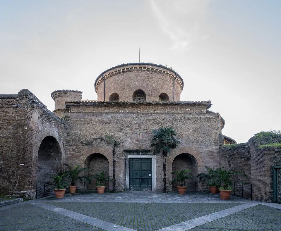 A round brick drum with arched windows rising behind a lower front of rough brick and stone, with a plain doorway between two blind arches, potted palms along the wall and a paved courtyard in front.