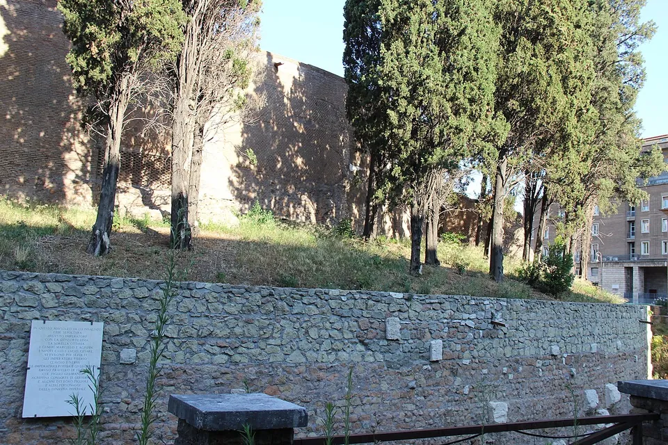 A high curved wall of brick and patchy stone rising behind cypresses on a grassy bank; below, a retaining wall of rough grey stones carries a white marble plaque with a long inscription in capitals.