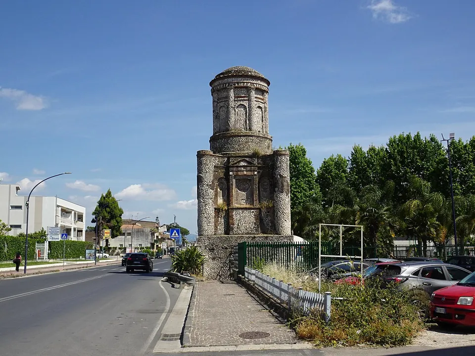 A two-storey Roman tomb rising at the edge of a main road, next to a pavement and a small car park, with apartment blocks, palm trees and street lamps around it under a blue sky.