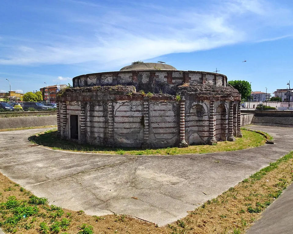 A low, round Roman building of two storeys standing in a sunken, paved enclosure ringed by a strip of grass. The lower storey has arched niches between pilasters banded in red brick, a narrower upper ring sits on top, and parked cars and modern buildings show beyond the rim of the enclosure.