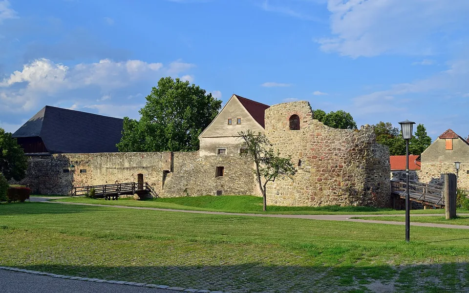 A long stretch of old stone wall with a rounded tower, running behind a wide lawn with a lamp post and a young tree, houses with dark roofs beyond it.