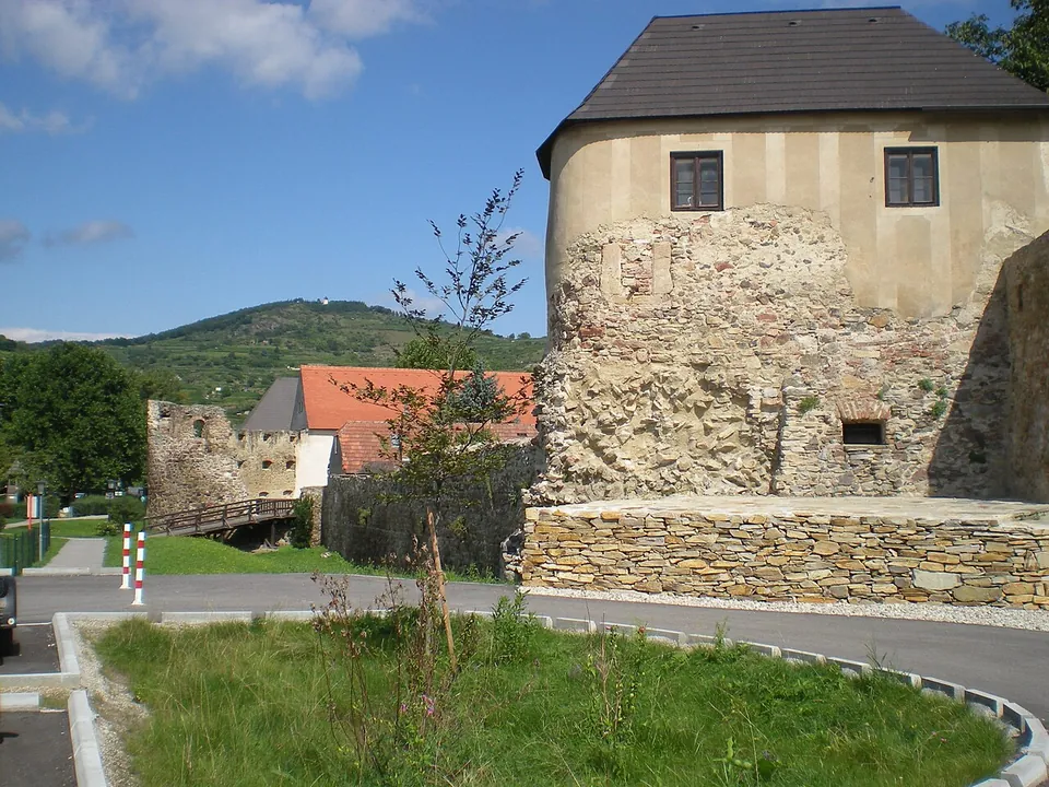 A broad rounded tower of rubble stone, plastered above and carrying a later storey with windows and a dark roof, beside a road; behind it a low wall and a footbridge lead to a ruined tower, with red-roofed houses and vineyard hills beyond.