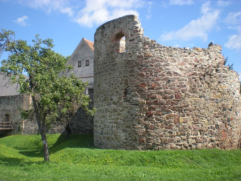 A tall, round-fronted tower of rough rubble stone with a small brick-arched opening near the top, standing on a lawn beside a fruit tree, with a stretch of wall and an old house behind.