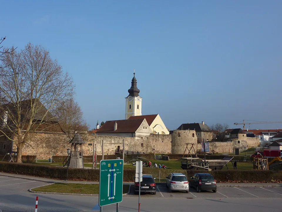 A car park with a few cars and a green road sign in the foreground, and beyond it a low old wall with a round tower, a church tower and other roofs.