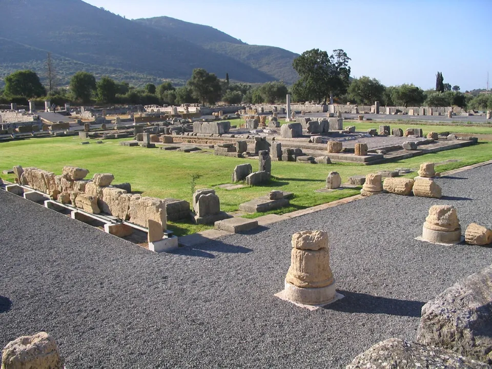 A wide green courtyard surrounded by rows of column bases and scattered stone blocks on gravel paths, with more ruins and trees beyond and a mountain behind.