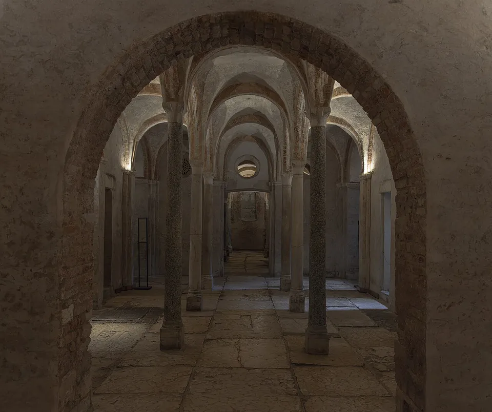 Looking through a round brick arch down the length of a low vaulted crypt, its stone columns running in rows towards a lit far wall, the floor laid with large worn stone slabs of uneven size.