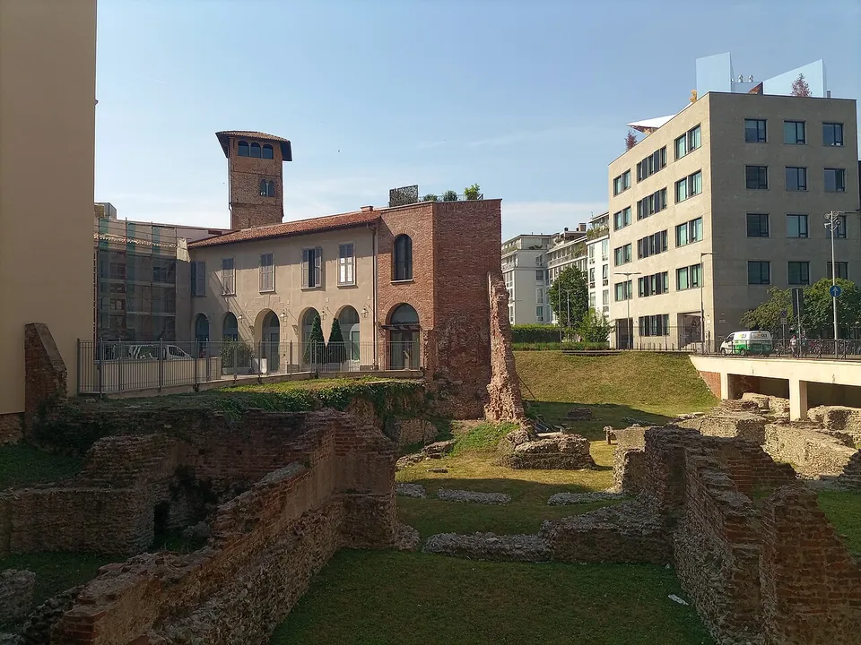 A green plot of brick and rubble walls standing a little above the grass, a railing along its far side, with an old brick house and tower and modern office blocks behind it.